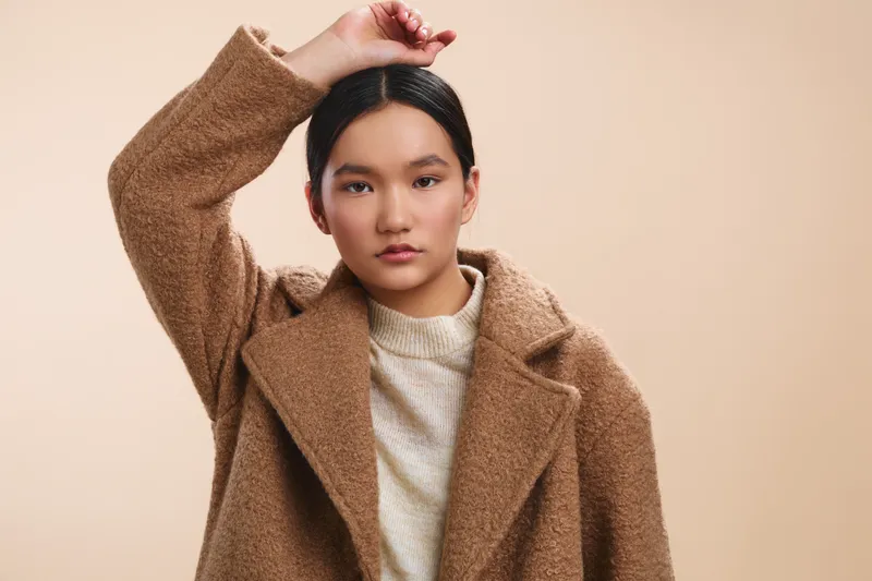 asian woman posing in a warm beige studio, wearing a cozy wool coat and light sweater, gazing at the camera with calm confidence, creating a modern, minimalist portrait that exudes elegance and ease.