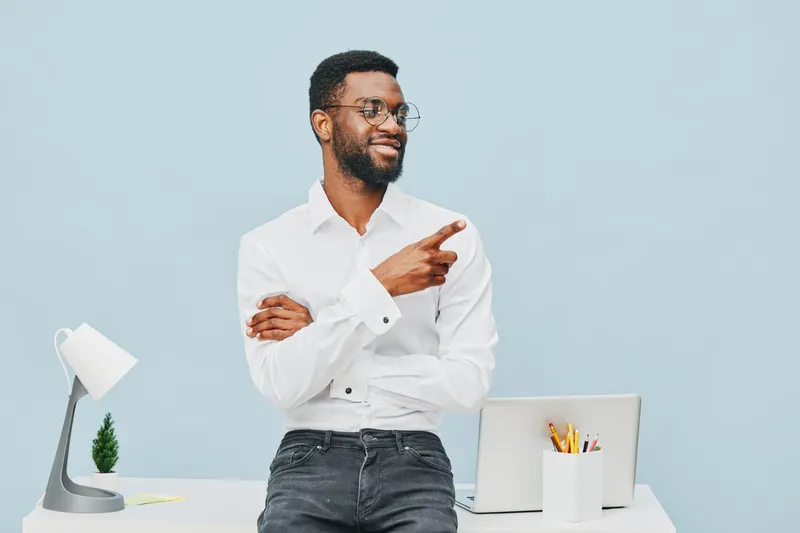 Smiling man with glasses wearing white shirt standing at modern office desk, pointing finger towards right side, adults workspace concept, professional business environment, positive mood, casual styl