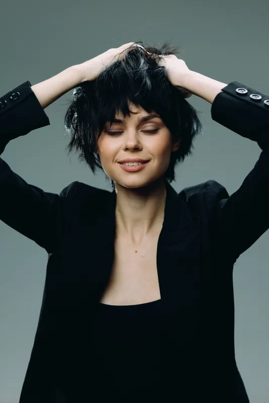 smiling woman with short black hair wearing a black blazer with closed eyes and hands in hair against gray background