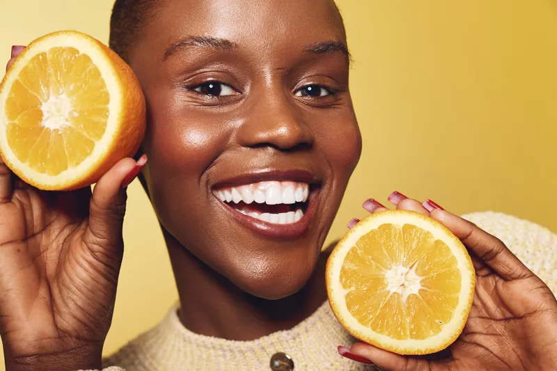 Smiling black woman with dark skin holds two halves of a fresh orange close to her face. Bright yellow background and warm lighting emphasize healthy skin and natural beauty concept.