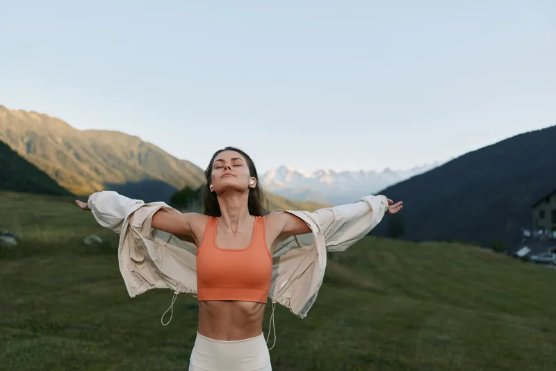 A carefree woman stands outdoors with arms wide, wearing athletic gear in a scenic mountain meadow. She looks joyful and liberated, savoring fresh air and wide horizons.
