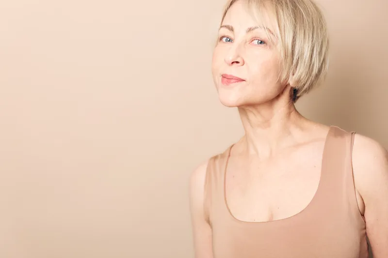 Confident mature woman with short blonde hair and natural makeup poses against beige background wearing sleeveless top. Concept of aging beauty, self acceptance, and simplicity.