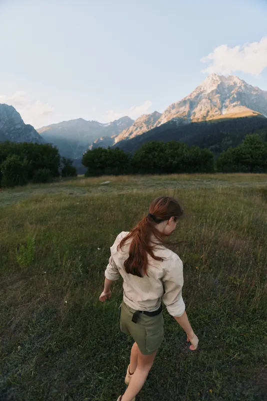 Woman lifestyle on a meadow with distant mountains, back view, dressed in casual shirt and shorts, exploring nature and enjoying peaceful outdoor scenery during golden hour.