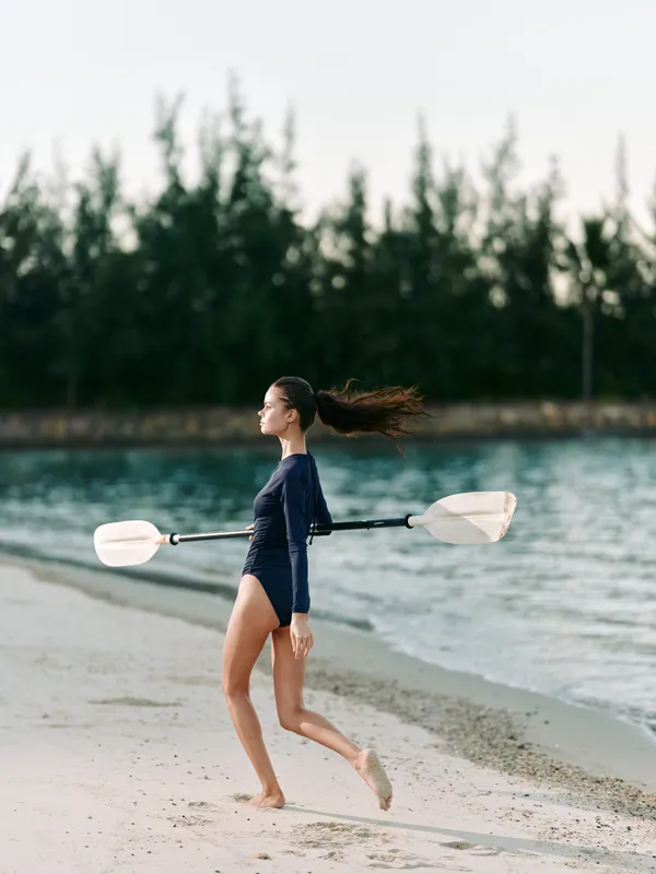 Young paddleboarder runs along a sandy shoreline, carrying a double paddle across her shoulders while water glints nearby and distant pines frame a moment of motion and freedom.