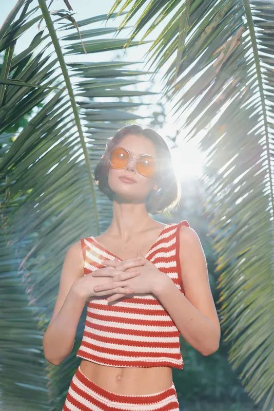 A stylish woman wearing a red and white striped crop top and sunglasses stands in a lush tropical setting, basking in warm sunlight and exuding carefree confidence.