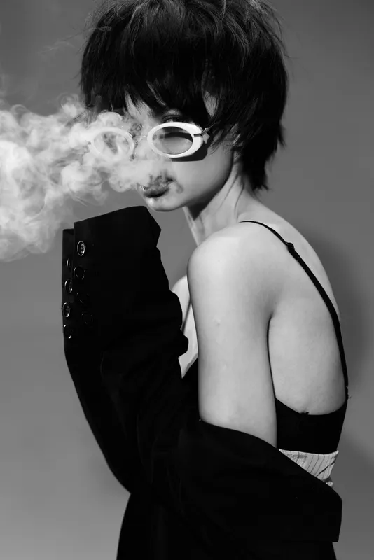 Black and white portrait of a young woman with dark hair wearing sunglasses, blowing smoke, expressing mystery and cool style.