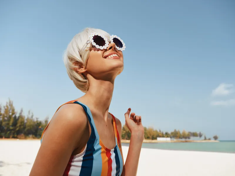 Sunny beach scene featuring a smiling woman with short blonde hair, round sunglasses, and a striped swimsuit, enjoying warm light and carefree summer vibes by the sea.