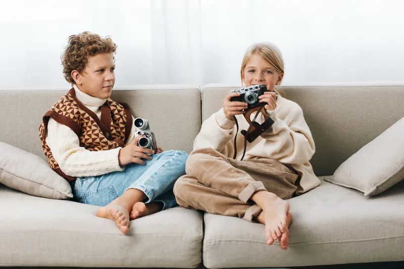 Two children sit on a light sofa, each holding a vintage style camera, absorbed in playful photography as natural light fills a calm, cozy living room.