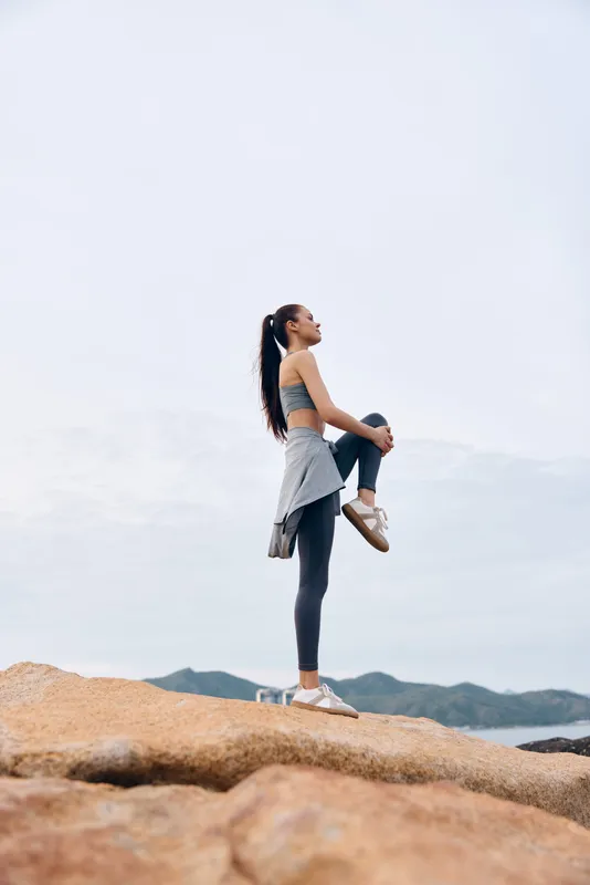 Young athletic woman stretching on rocky terrain with mountains in the background, wearing grey workout clothes, expressing determination and focus