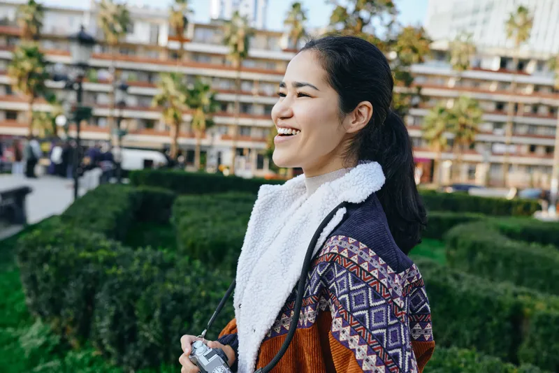 Smiling woman in a knit scarf enjoys an outdoor park setting with greenery and modern buildings in the background, showcasing casual fashion and relaxed mood during daytime.