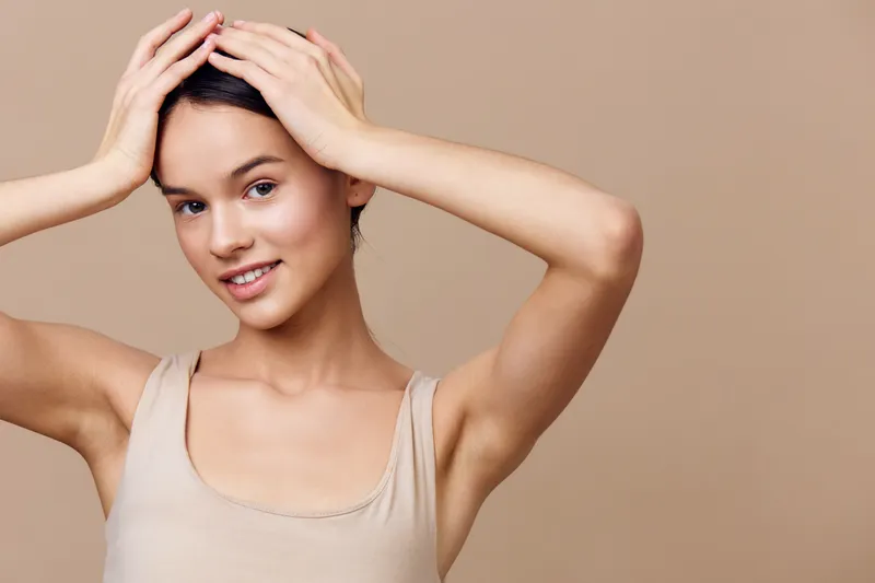 Young woman with smooth skin holding her head, showcasing healthy beauty and skincare, against a neutral beige background, ideal for wellness and beauty themes
