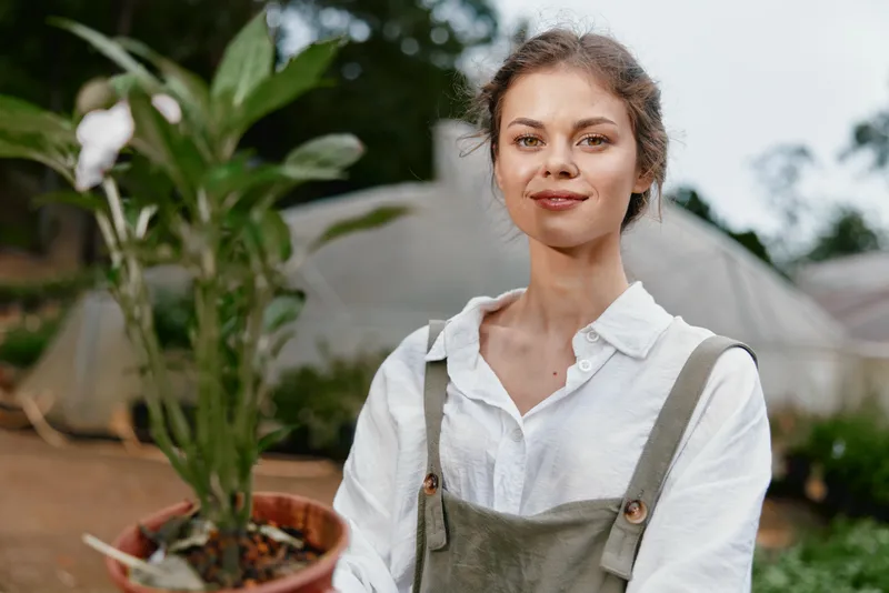 Young woman with a potted plant in casual gardening attire smiles gently in a greenhouse setting, showcasing a connection with nature and sustainability