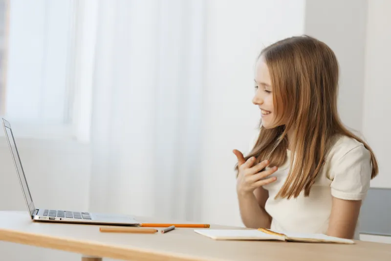Young girl engaging in a video call, showcasing excitement and happiness while sitting at a desk with a laptop, bright natural lighting enhances the cheerful atmosphere