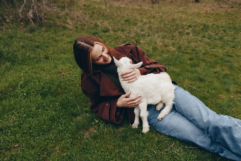 Woman relaxing on grass embraces a playful goat kid in a rural setting, a tender moment of affection, nature, and outdoor lifestyle, with soft light creating a warm, peaceful atmosphere