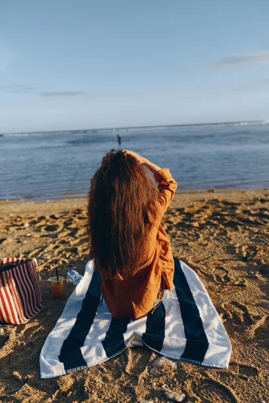 Woman on beach, sea horizon behind, sunlight on curly hair, striped towel on sand, relaxed posture, warm summer mood, gentle breeze, natural colors, quiet escape