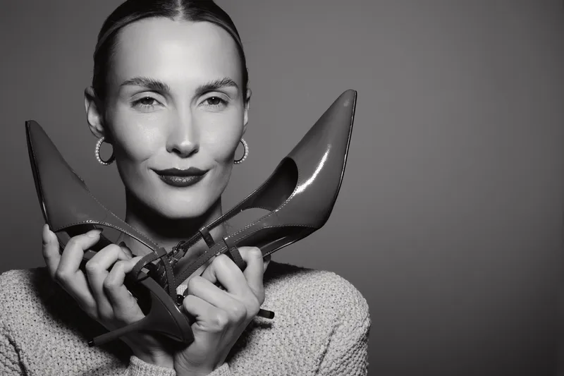 White woman with light skin holds red high heels near face looking confidently at camera. Fashion concept with elegant style and beauty in studio lighting.