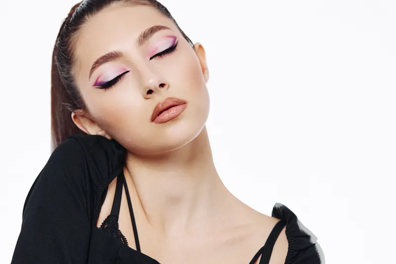 Young woman with brown hair and vibrant purple eyeshadow poses with closed eyes wearing lipstick and black fashion outfit against a plain white background. Concept beauty creative fashion.