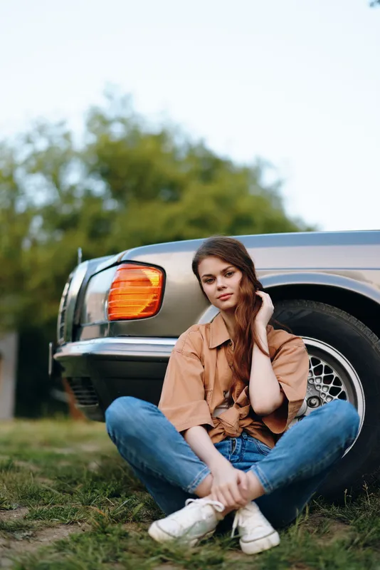 Young woman with long hair sitting by a vintage car in casual attire, conveying a relaxed summer vibe with a gentle smile against a green background.