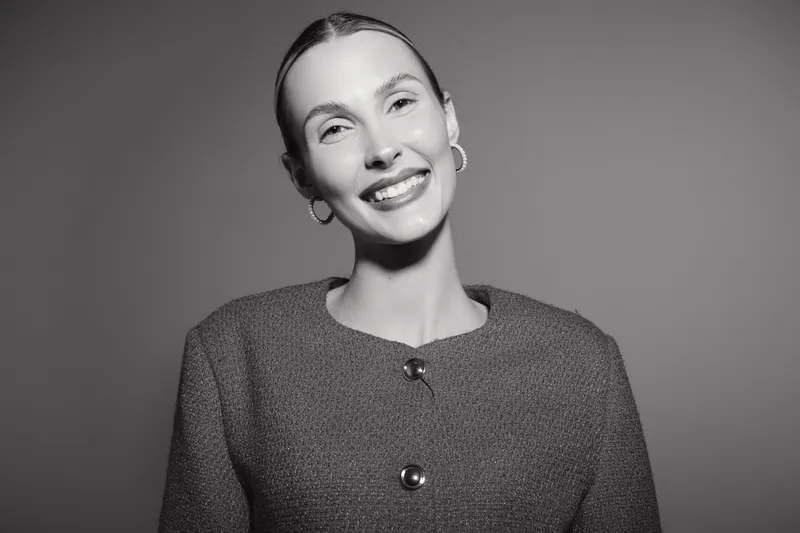 Smiling young white woman with light skin and slicked back hair wears a textured jacket, posing confidently in a studio. Concept of happiness, fashion, and portrait photography.