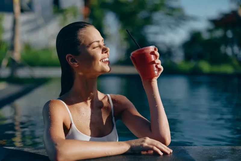 A woman lounges by a pool with a red cup, smiling in bright sunlight, wearing a white swimsuit. A light, casual lifestyle scene focused on relaxation and hydration.