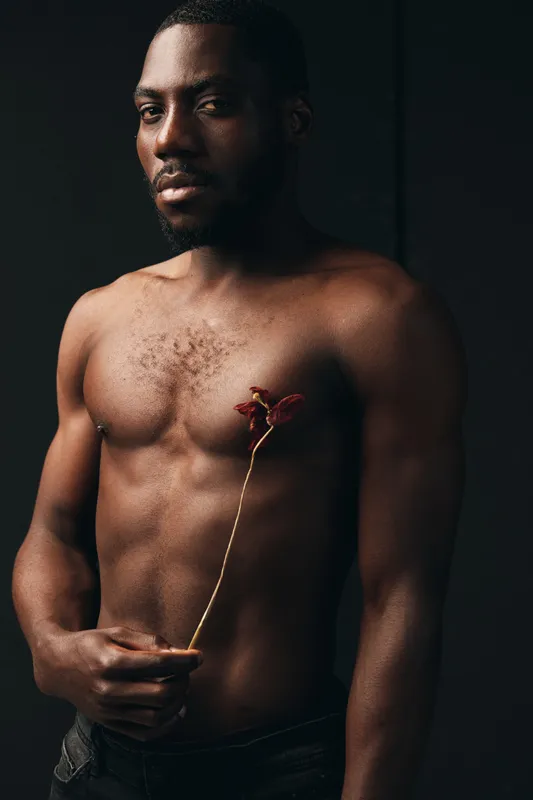 African man with dark skin and muscular body holding a dried flower in creative conceptual visual style. Portrait with moody lighting and artistic expression on black background.