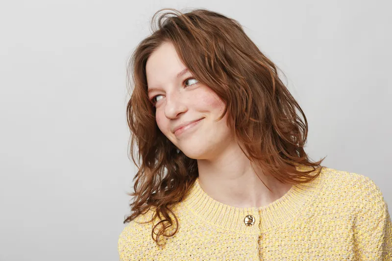 Young woman with curly brown hair wearing yellow sweater looks sideways with a slight smile on light grey background. Concept of casual style, natural beauty, and positive mood.