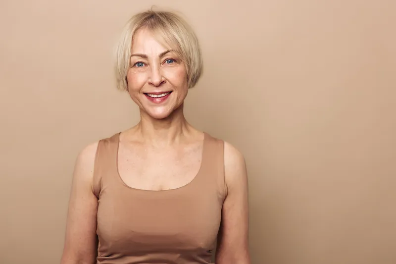 Smiling mature woman with short blonde hair wearing beige tank top poses against neutral background. Concept of happiness, aging, confidence, and natural beauty in studio portrait.
