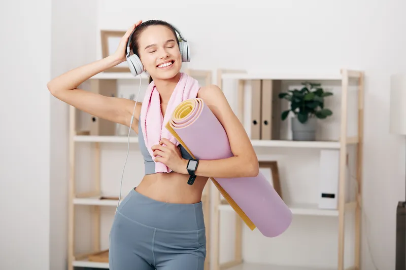 Active woman fitness scene at a bright home gym, wearing headphones, holding a rolled yoga mat and towel, smiling during a focused, upbeat workout session.