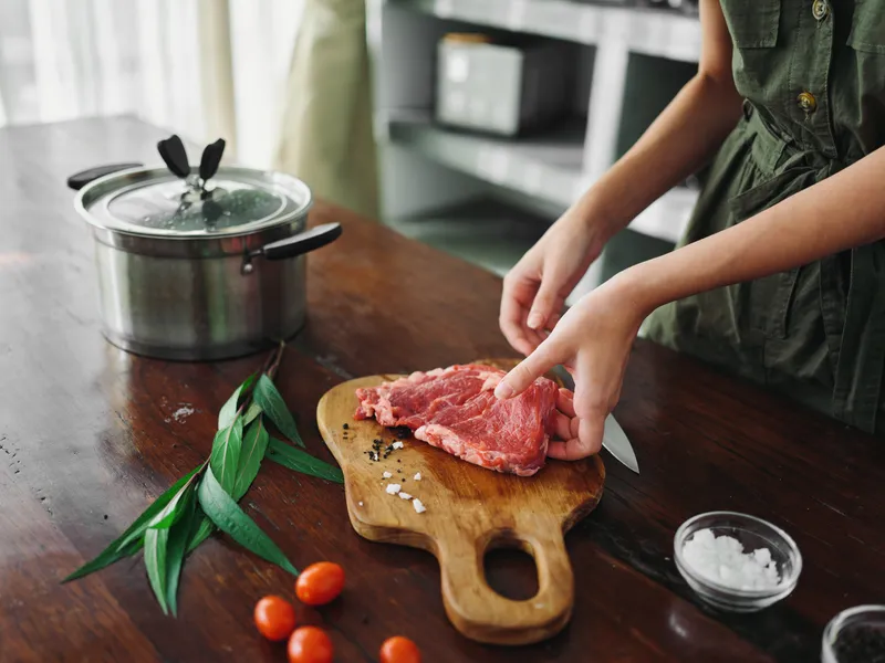 Женщина на кухне готовит ужин из мяса, овощей и специй на деревянном столе на фоне стильной кухни