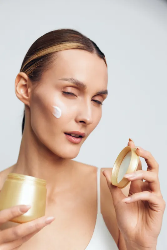 Young woman with clear skin applying white face cream on cheek. Beauty skincare routine in bright studio light. Concept of moisturizing, self-care, and healthy skin.