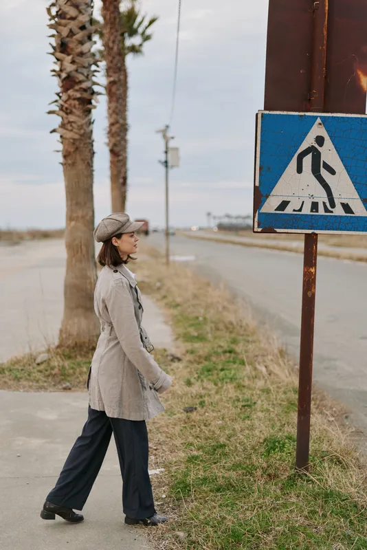 Woman in cozy sweater and hat walking near pedestrian crossing sign on rural roadside with palm trees under cloudy sky. Concept of autumn fashion, casual style, and outdoor lifestyle.