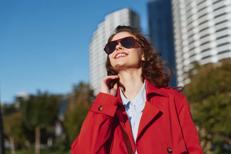 Happy woman outdoors wearing red coat and sunglasses smiling with closed eyes sunny day urban background modern buildings