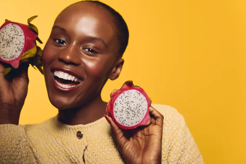 Happy African woman with dark skin holds two halves of dragon fruit, smiling brightly against yellow background. Concept of healthy lifestyle, tropical fruit, and natural beauty.