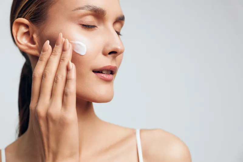 Young woman with clear skin applies moisturizing cream on her cheek with closed eyes in a calm skincare routine against a plain light background.