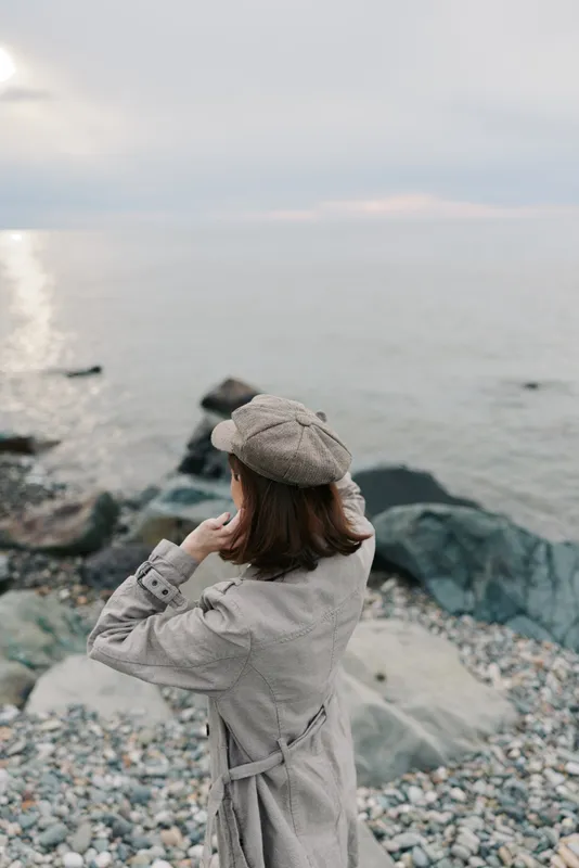 A woman in a grey coat and matching hat stands on a rocky beach looking out over a calm sea under a cloudy sky. Concept of solitude, contemplation, and autumn fashion.