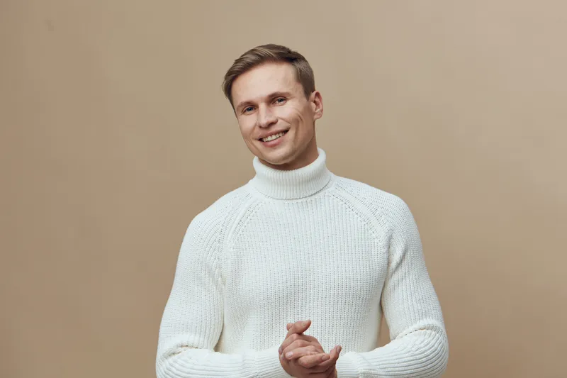 smiling young caucasian man wear white knitted turtleneck sweater isolated on brown background indoor portrait mental health gadgets concept