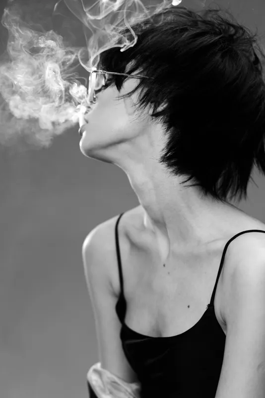 Black and white portrait of a young woman blowing smoke in a stylish black dress with short hair.