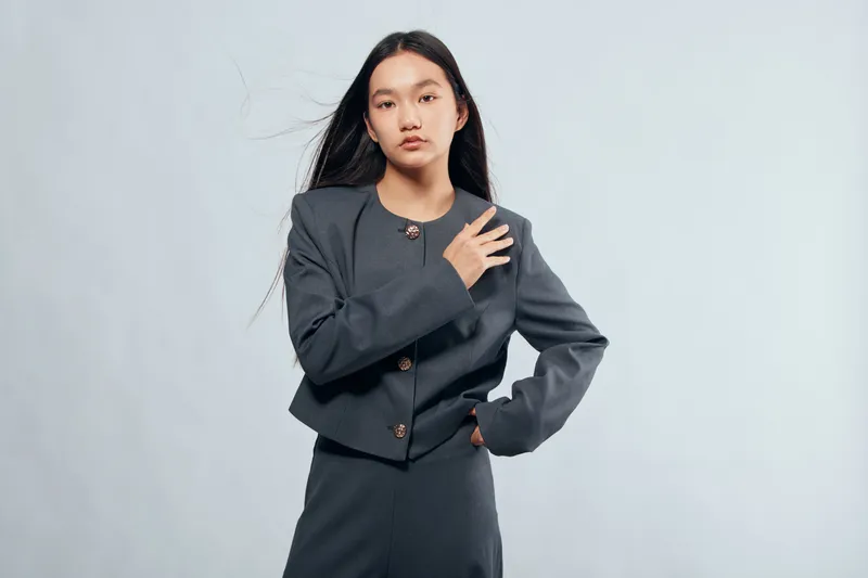 Young woman standing with hand on chest wearing dark gray jacket and skirt, long black hair, serious expression, neutral background portrait for fashion or business concept.