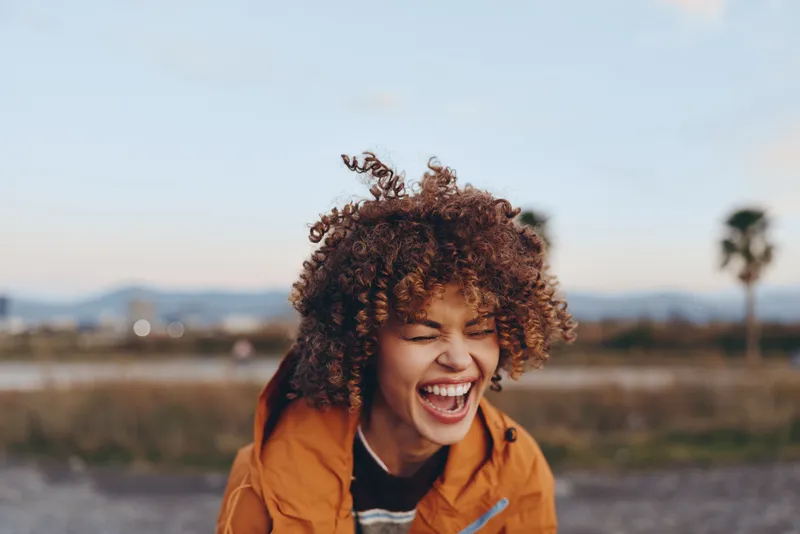 Woman lifestyle outdoors sunset smiling curly hair portrait of a laughing woman in an orange jacket, warm sky over a field, soft light, background blur with distant trees and open nature