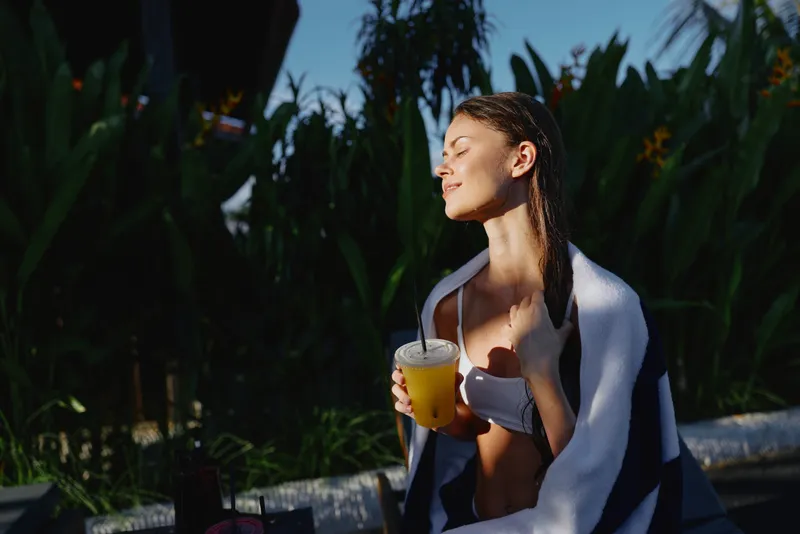 A young woman enjoys sunlight while holding a glass of orange juice wrapped in a blanket outdoors. The tropical plants create a relaxing lifestyle atmosphere.