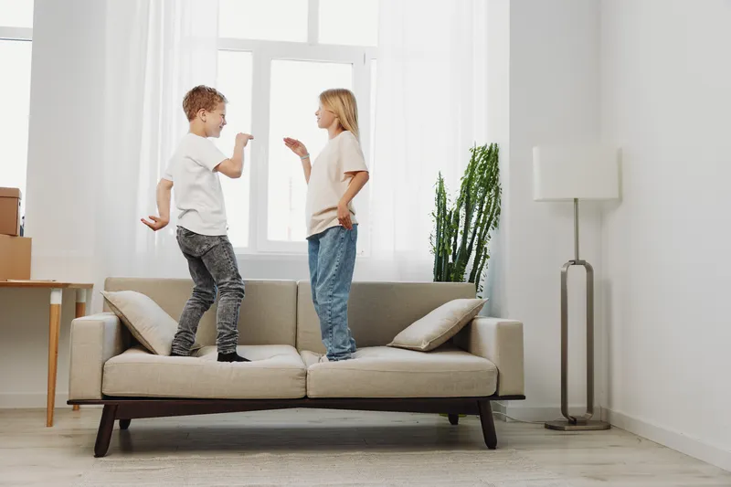 Two children standing on sofa in bright living room playfully interacting, casual clothes, natural light, modern minimal interior with plant and lamp.