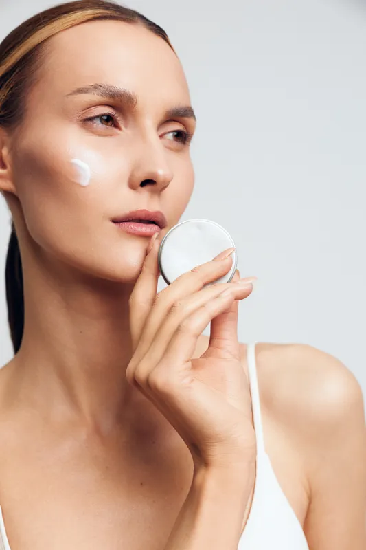 Young woman with clean skin applying moisturizer cream on cheek using cotton pad, gentle skincare routine, natural beauty, soft lighting, minimal makeup, white background