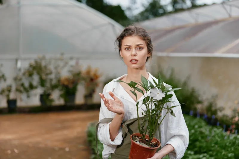 Young woman surprised in a greenhouse holding a potted plant, expressing curiosity and concern about gardening and nature during springtime