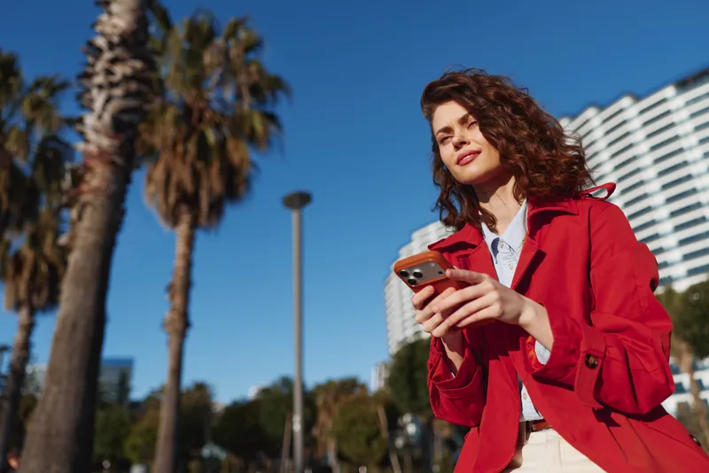 Young woman outdoors in a red coat holds a smartphone, smiling, under bright sun with palm trees and city buildings in the background