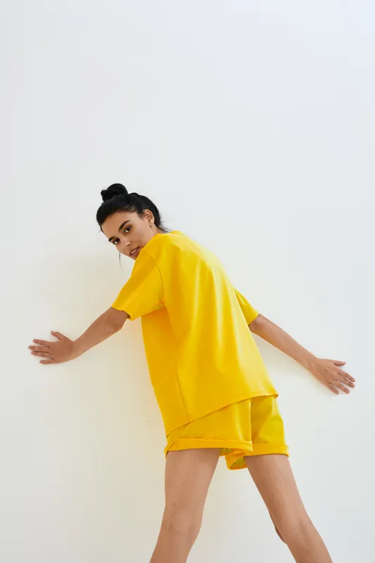young woman in yellow outfit leaning against white wall with casual posture and natural lighting, contemporary style, minimalistic background in bright tones