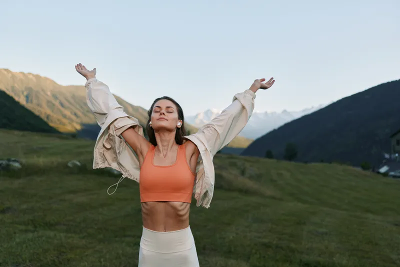 A fit woman stands in a mountain meadow with arms raised, enjoying fresh air and sunshine. She wears an orange sports top and light jacket, radiating confidence, energy, and a healthy outdoor