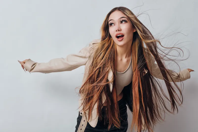 Young woman with long brown hair and dreadlocks wearing stylish beige jacket and light top posing with hair flying on white background in studio.