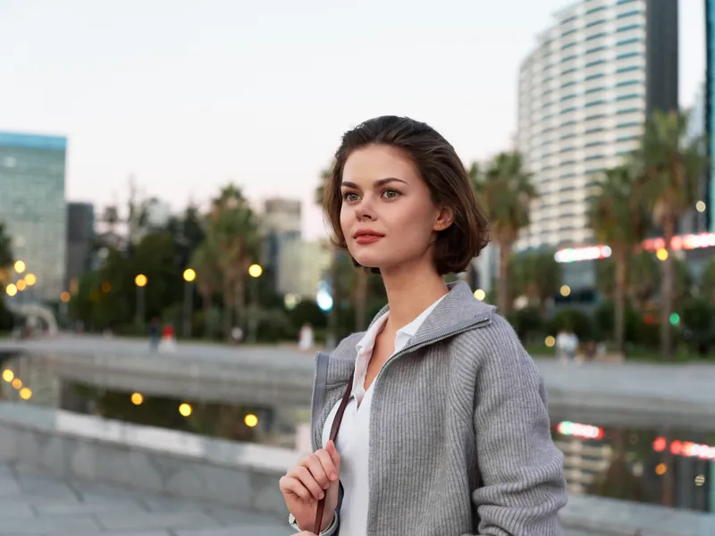 A poised professional woman stands outdoors in a modern city setting, wearing a light coat and white shirt, looking forward with calm confidence and poise.