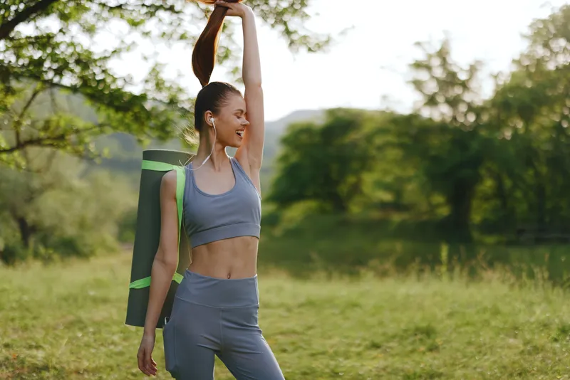 Young athletic woman stretching in a green park, wearing a grey sports outfit and holding a yoga mat, embodying fitness and health