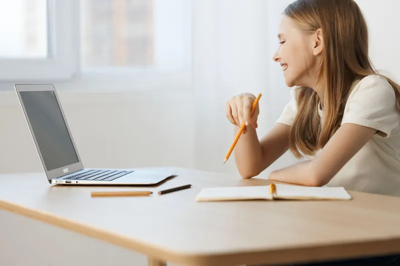 Smiling young woman studying online with a laptop, showcasing a positive learning environment, bright sunlight and modern workspace aesthetic